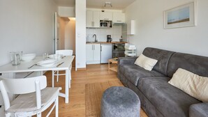 A modern living room with a grey sofa, white dining table, and chairs. A kitchen with white cabinets and appliances is visible in the background. - Un Balcon sur la Mer - Appt à 30m de la plage (Saint-Aubin-sur-Mer)