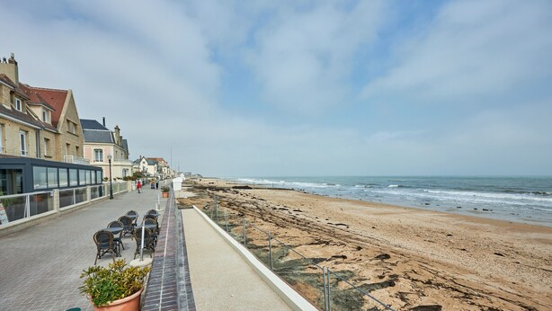 A seaside promenade with buildings, a beach, and a row of outdoor tables and chairs. - Un Balcon sur la Mer - Appt à 30m de la plage (Saint-Aubin-sur-Mer)