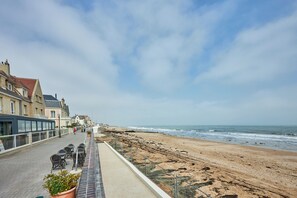 A seaside promenade with buildings, a beach, and a row of outdoor tables and chairs. - Un Balcon sur la Mer - Appt à 30m de la plage (Saint-Aubin-sur-Mer)
