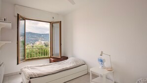 A bedroom with a bed, a bedside table with a lamp, a window with a view of buildings and hills, and a ceiling fan. - Alivi - Appt climatisé avec vue mer et montagne (L'Île-Rousse)