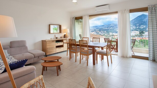 A modern living room with a sofa, dining table, chairs, and a view of a mountainous landscape through large windows. - Alivi - Appt climatisé avec vue mer et montagne (L'Île-Rousse)