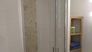 Een douchegedeelte met een glazen deur, betegelde wand, een houten plank met een doos en een prullenbak.