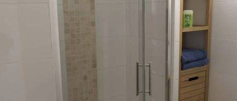 Un espacio de ducha con puerta de cristal, pared de azulejos, repisa de madera con una caja y un cubo de basura.