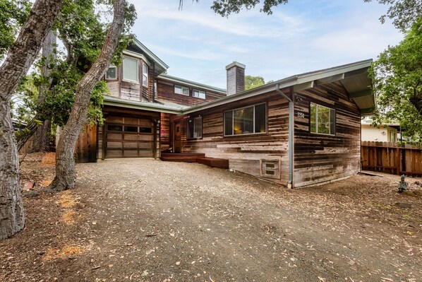 A two-story wooden house with a garage, surrounded by trees and a gravel driveway.