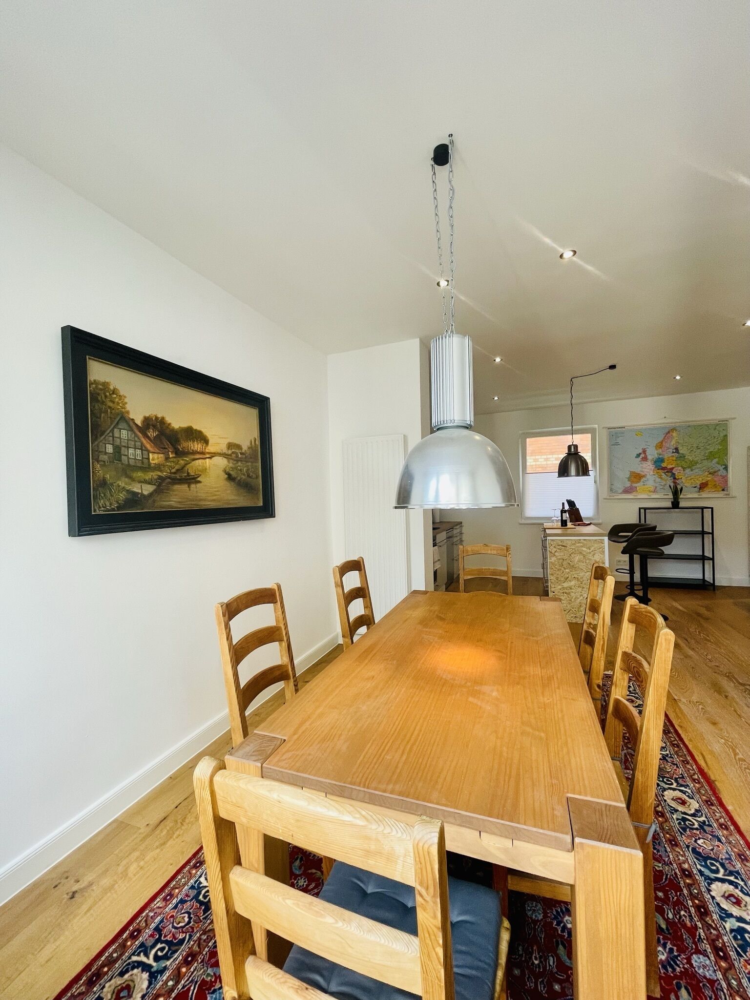 Un comedor con una mesa y sillas de madera, una lámpara colgante, un cuadro enmarcado en la pared y una cocina al fondo.