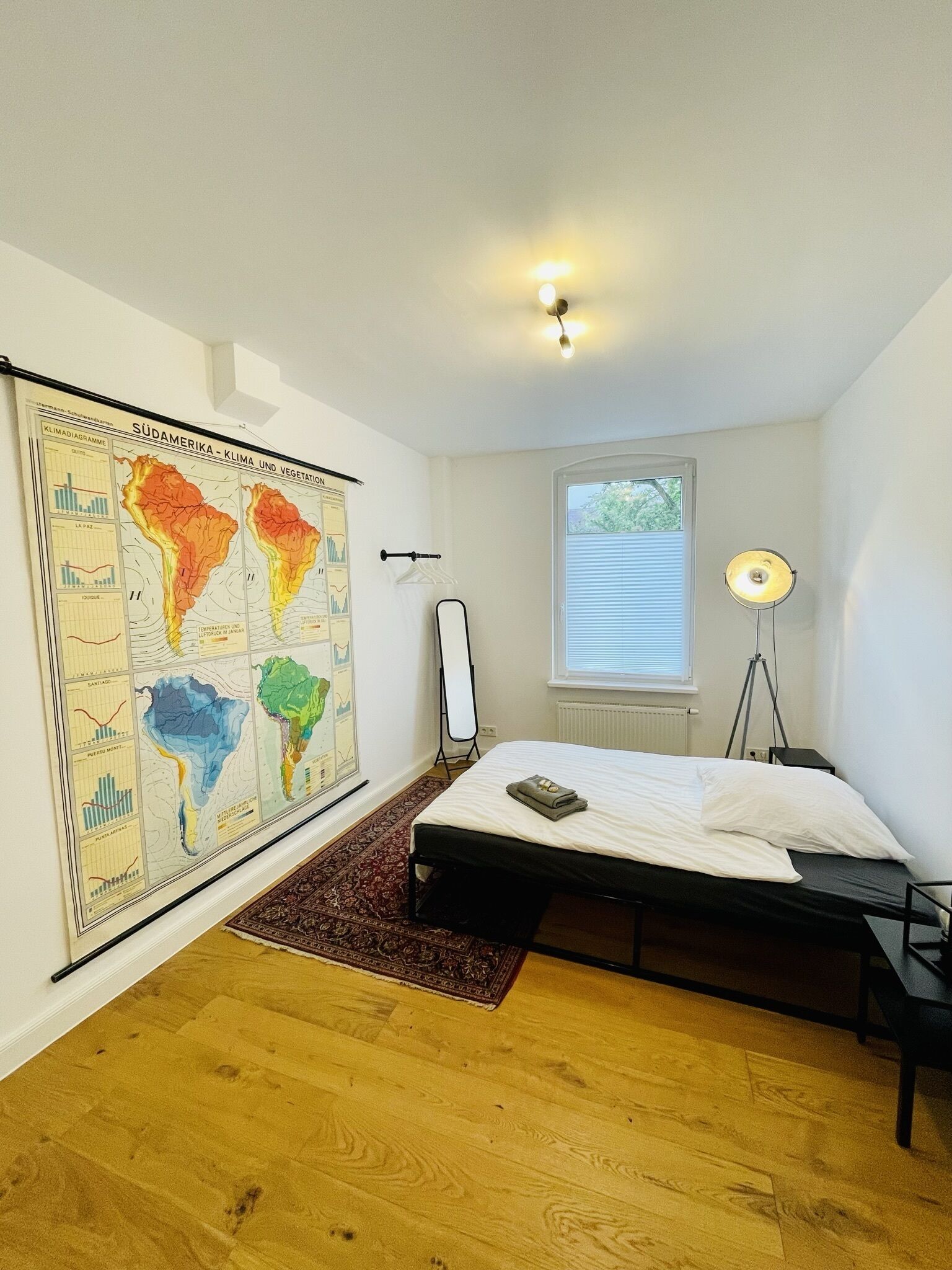 A bedroom with a bed, a map on the wall, a standing lamp, and a window.