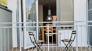 Un balcón con una mesa y sillas, una lámpara colgante, y vista del interior a través de una puerta abierta.
