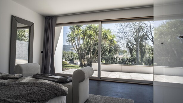 A modern bedroom with a large sliding glass door leading to a balcony with a view of palm trees and a garden. - Casa da Magnólia (Briteiros (Santo Estêvão))