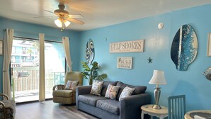 Un soggiorno con una parete blu, un divano grigio, un tavolino e una sedia. C'è una ventola a soffitto, una finestra con tende e un cartello con la scritta "Gulf Shores Alabama Benvenuti al Beach House".