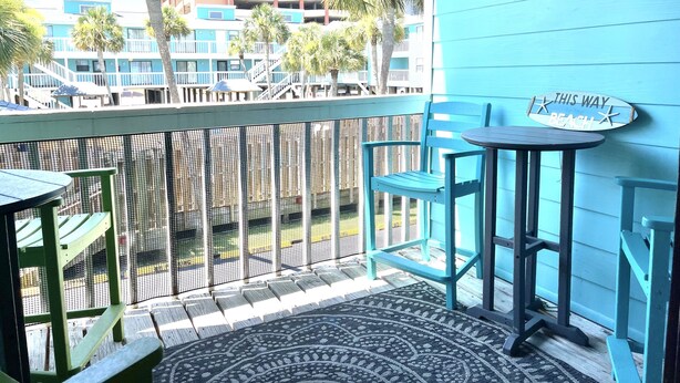 Un balcone con vista su un edificio, una sedia, un tavolino e un cartello con su scritto 'THIS WAY TO THE BEACH'.