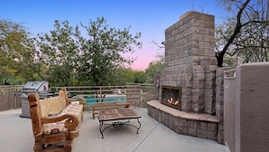 Un patio exterior con chimenea de piedra, banca de madera, mesa y piscina al fondo.