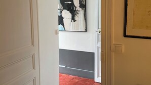 Een hal met een witte deur, een vloer van rode tegels en twee kunstwerken in lijsten aan de muur.