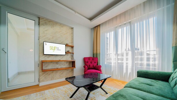 A modern living room with a green sofa, a red armchair, and a black coffee table. There is a TV mounted on the wall and a window with sheer curtains.