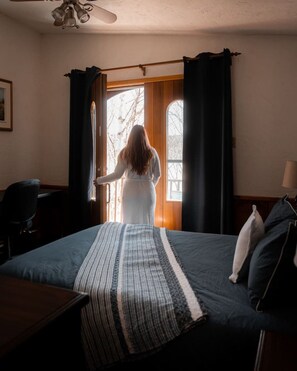 Una habitación con cama, una silla, un ventanal con cortinas y un ventilador de techo.