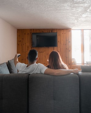 Una pareja sentada en un sofá gris frente a un televisor de pantalla plana montado en una pared de madera.