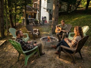 A group of people sitting around a fire pit in an outdoor setting with chairs and a deck in the background.