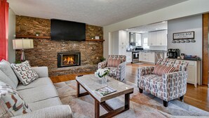 A living room with a stone fireplace, a flat-screen TV, a coffee bar, and a kitchen in the background. - Aiken Rd - 6 BR - hot tub and game room (Asheville)