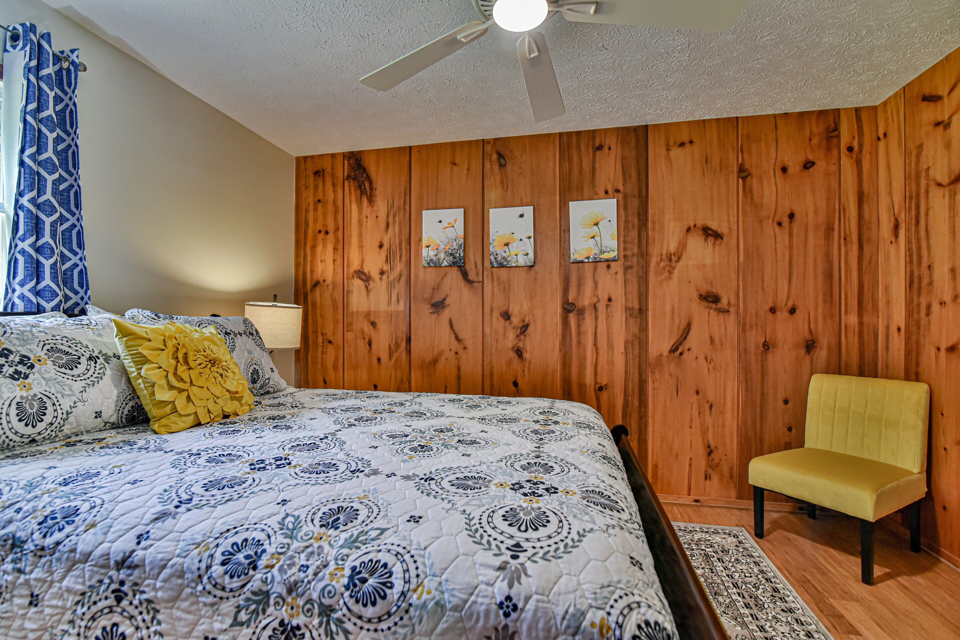 Quarto com parede de painel de madeira, cama com colcha floral, uma cadeira amarela e um ventilador de teto.