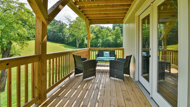 A wooden deck with a covered patio area, featuring two wicker chairs and a table, overlooking a grassy landscape. - Aiken Rd - 6 BR - hot tub and game room (Asheville)