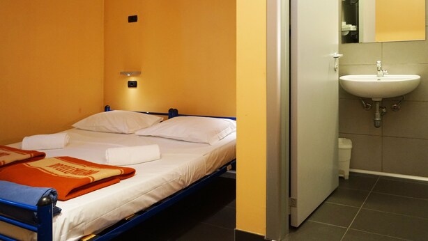 A single bed with white linens and a blue metal frame, a white wall with a small light fixture, and an open door leading to a bathroom with a white sink and tiled walls. - Hi! Ostello Milano (Milan)