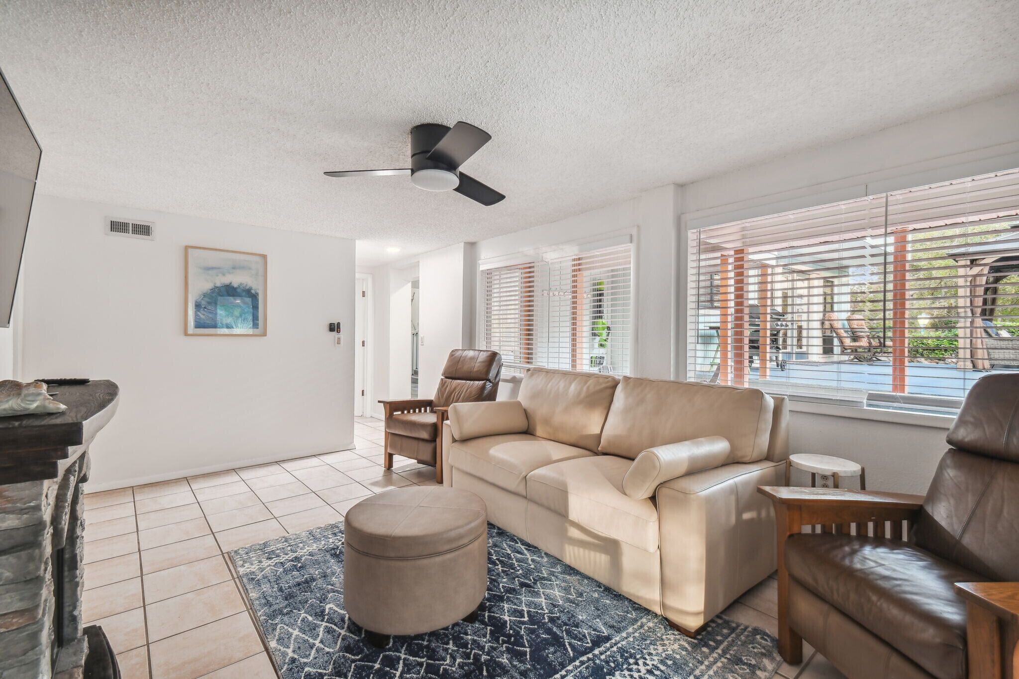 A modern living room with a beige leather sofa, a brown leather chair, a small round ottoman, and a ceiling fan.