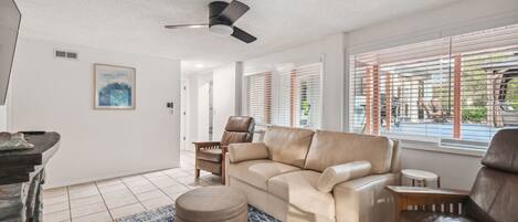 A modern living room with a beige leather sofa, a brown leather chair, a small round ottoman, and a ceiling fan.