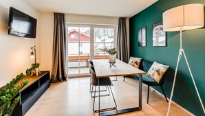 A modern living room with a wooden dining table, black chairs, a TV mounted on the wall, and a green accent wall. - VYLD: Owl's nest (Bad Hindelang)
