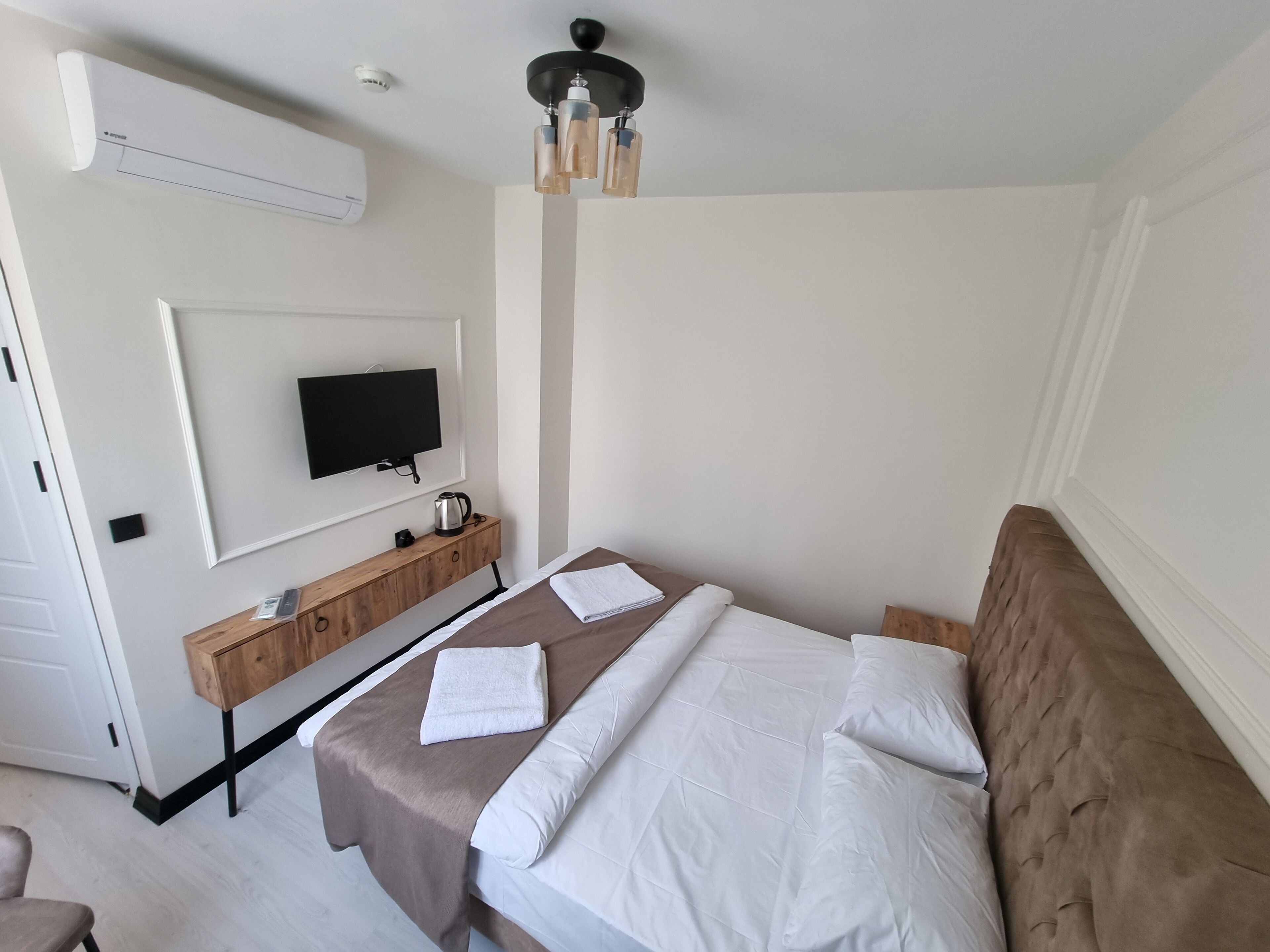 A hotel room with a bed, a TV mounted on the wall, a wooden side table with a kettle, and a ceiling fan.