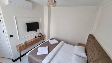 A hotel room with a bed, a TV mounted on the wall, a wooden side table with a kettle, and a ceiling fan.