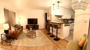 Una sala moderna con un sofá, una mesa de centro y taburetes de bar. Un televisor está montado en la pared, y se ve una cocina al fondo.