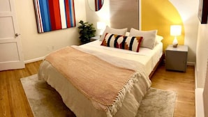 Un dormitorio con cama, mesita de noche con lámpara, espejo y un cuadro abstracto de colores en la pared.