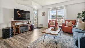 A modern living room with a flat-screen TV, leather sofas, a wooden coffee table, and a large window offering a view of the outdoors. - Lakeside Lair | Pool | Hot Tub | A/C (Invermere)