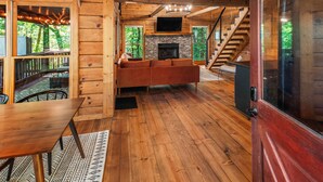 A wooden cabin interior with a stone fireplace, a wooden staircase, and a large window overlooking a forest. - The Maple Bear - Amazing Mountain Views + Hot Tub (Blue Ridge)