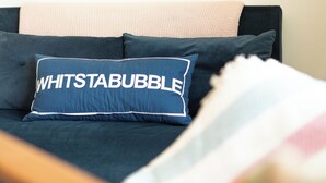 A bed with a dark blue pillow featuring the text 'WHITSTABUBBLE' and a light-colored headboard.