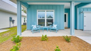 A covered porch with two blue chairs and a small table, a white garage door, and a house with blue siding. - Walk to Beach & Pool: Windmark Beach Home! (Port Saint Joe)