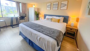 Ett hotellrum med en säng, sängbord, ett skrivbord och en stol. På väggen hänger tre inramade tavlor, det finns ett fönster med gardiner och utsikten utanför består av palmer och byggnader.