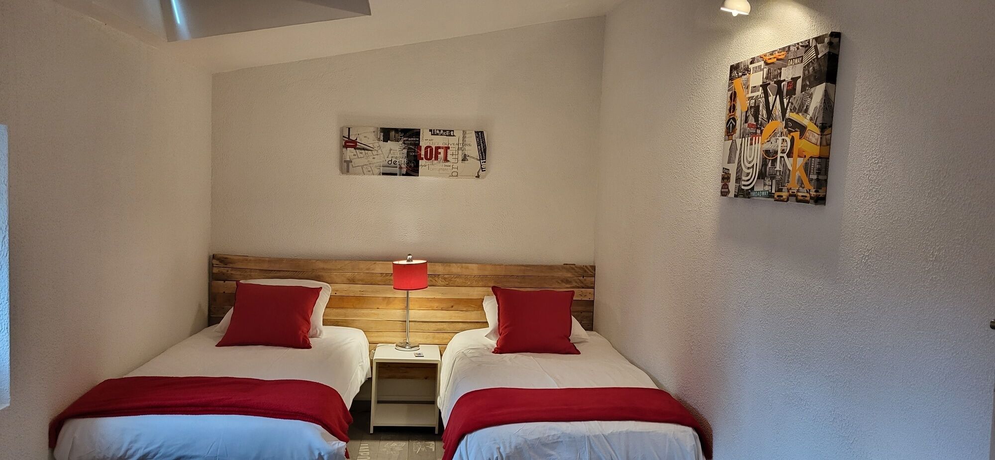 Ein Zimmer mit zwei Betten, jeweils mit roter Bettwäsche, einem Holzkopfende und einem Nachttisch mit Lampe. Über den Betten hängt ein Poster an der Wand.