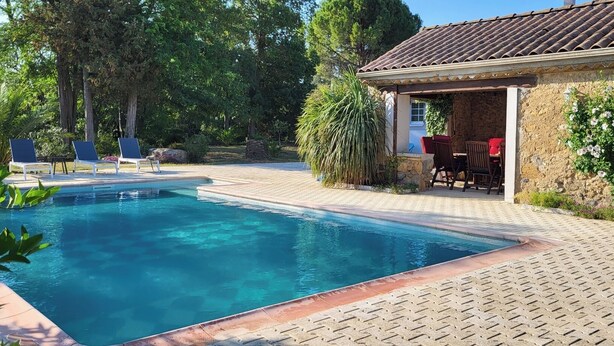 Une piscine dotée d’une terrasse carrelée, entourée de verdure et bordée d’un bâtiment en pierre avec un toit recouvert de tuiles.