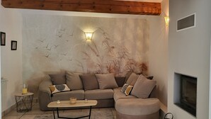 Un salon chaleureux avec un canapé, un pouf et une table basse, un mur orné d’une fresque représentant des oiseaux et une cheminée.