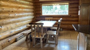 A log cabin interior with a wooden dining table and chairs, a window with a view of trees, and a kitchen area with wooden cabinets. - Stratton Ranch - Wyoming Getaway Cabin - 4 bed/2 bath WY Cabin! (Dubois)