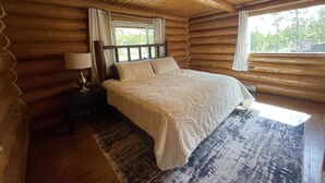 A log cabin bedroom with a wooden bed, a nightstand with a lamp, and a large window. - Stratton Ranch - Wyoming Getaway Cabin - 4 bed/2 bath WY Cabin! (Dubois)