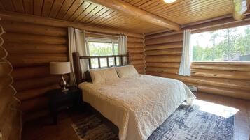A log cabin bedroom with a wooden bed, a nightstand with a lamp, and a large window.