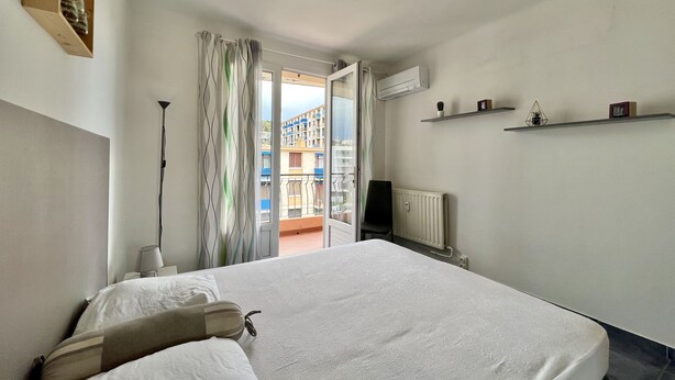 A hotel room with a bed, a chair, a radiator, a shelf with decorative items, and a view of a building outside. - Luminous cocoon near the beach (Saint-Raphaël)