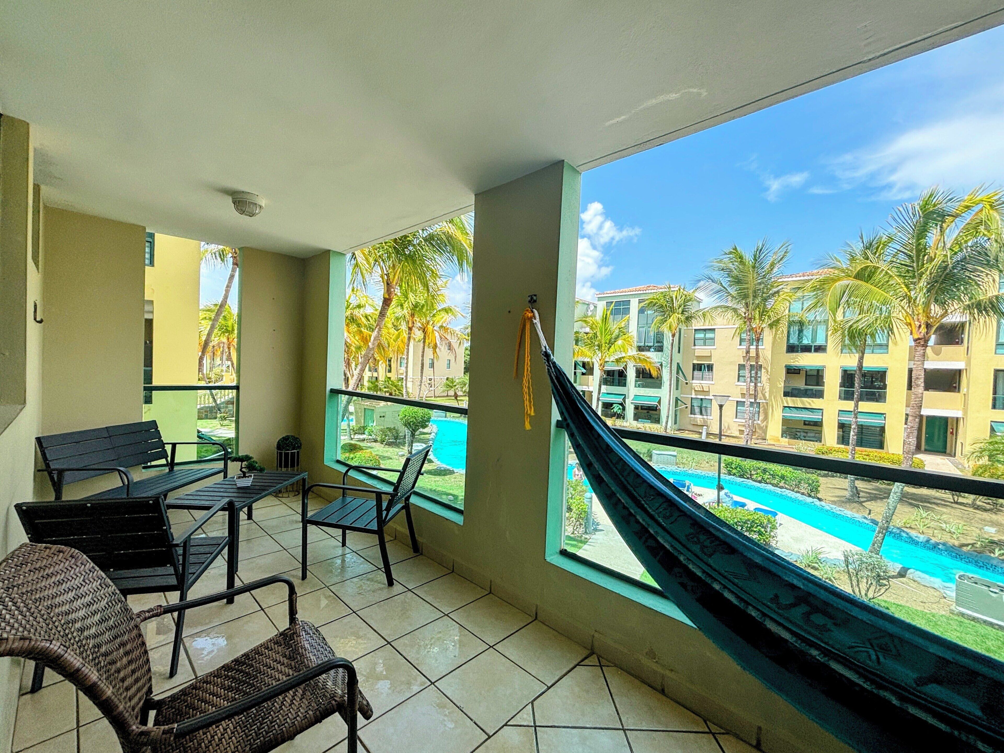 Un balcón con vista a una piscina, una hamaca y mobiliario exterior.