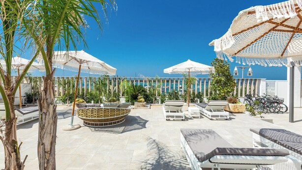 A sunlit outdoor patio with white wicker furniture, a round seating area, and umbrellas providing shade.