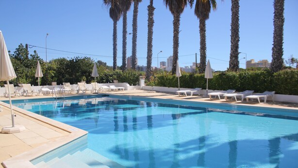 A swimming pool with lounge chairs and umbrellas, surrounded by palm trees and a cityscape in the background. - Villa Alcaide (Portimão)
