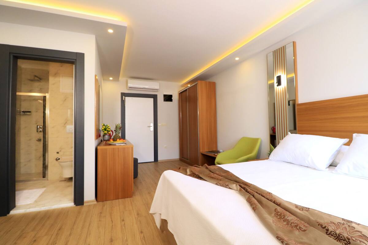 A modern hotel room with a wooden floor, a large bed with white linens, a wooden wardrobe, a green chair, and a small table with a vase.