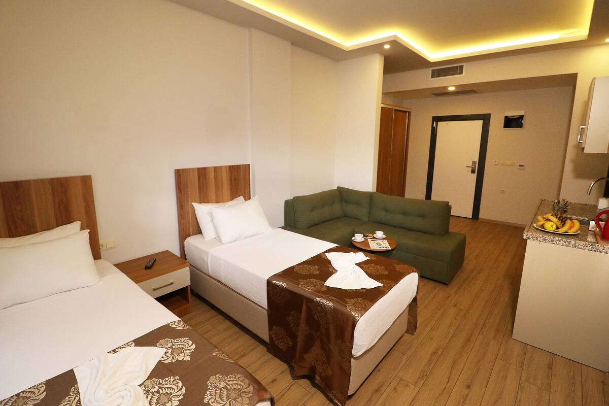 A hotel room with a bed, a sofa, a small table with a tray of fruit, and a kitchenette area.