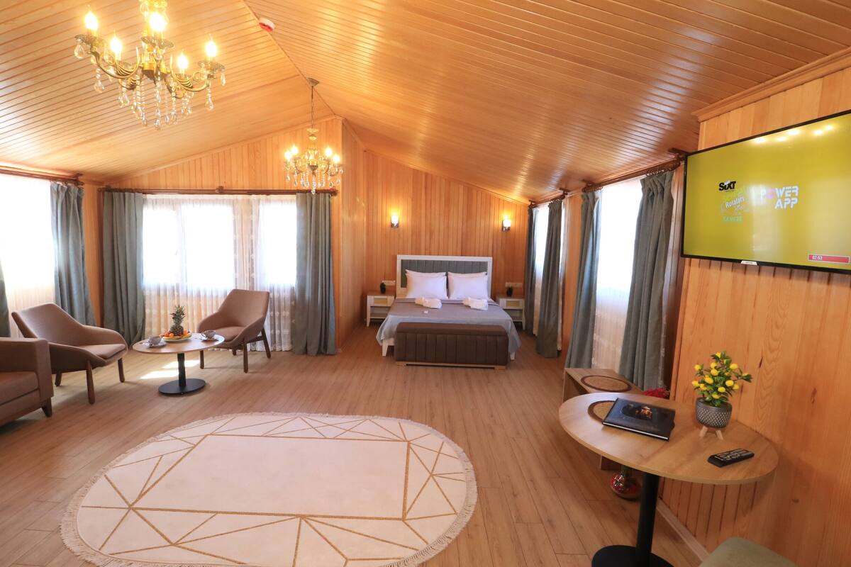 A hotel room with a bed, two armchairs, a round coffee table, a TV, and a wooden ceiling.