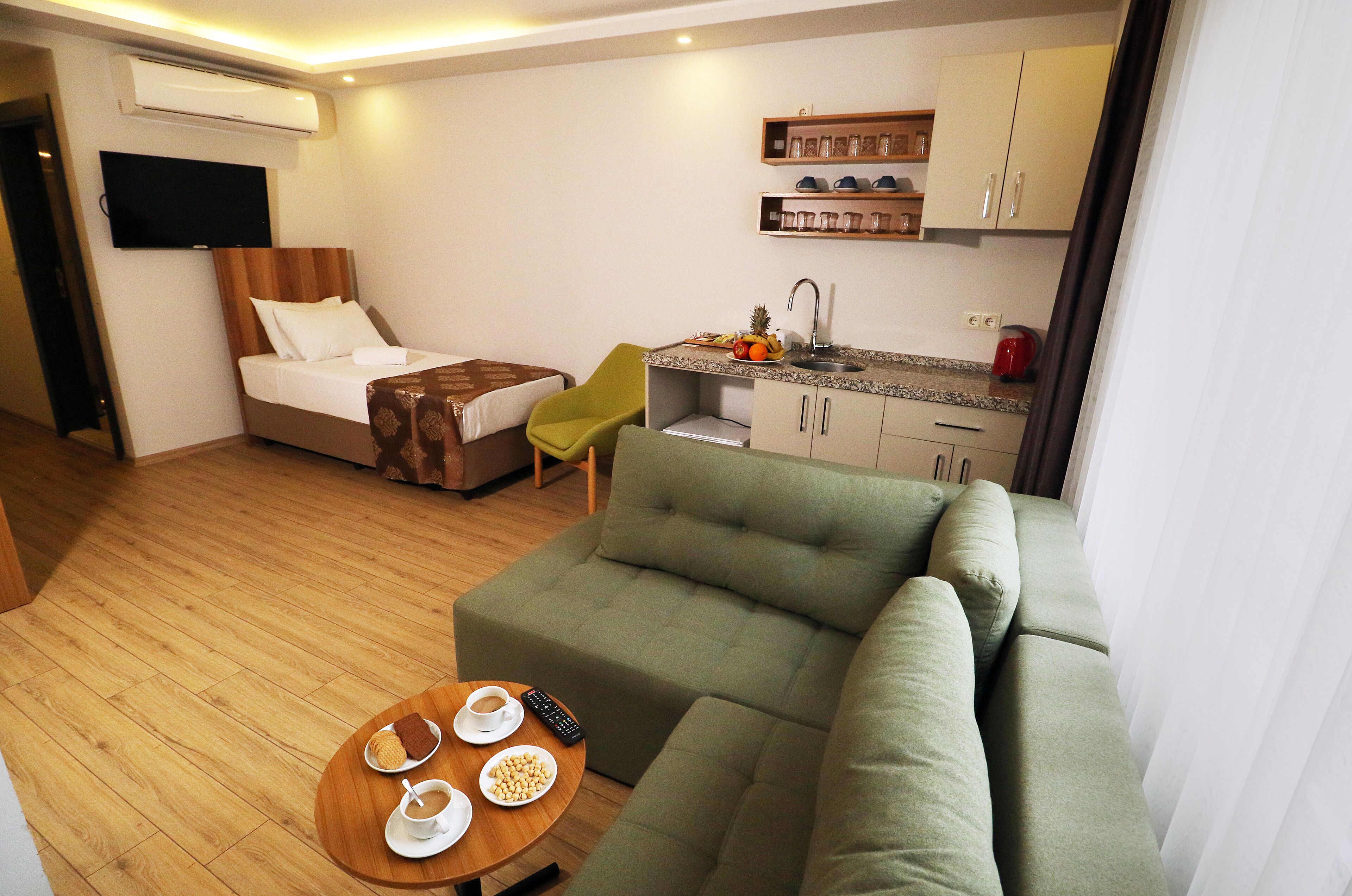 A hotel room with a bed, a green sofa, a small kitchen area, and a TV mounted on the wall.